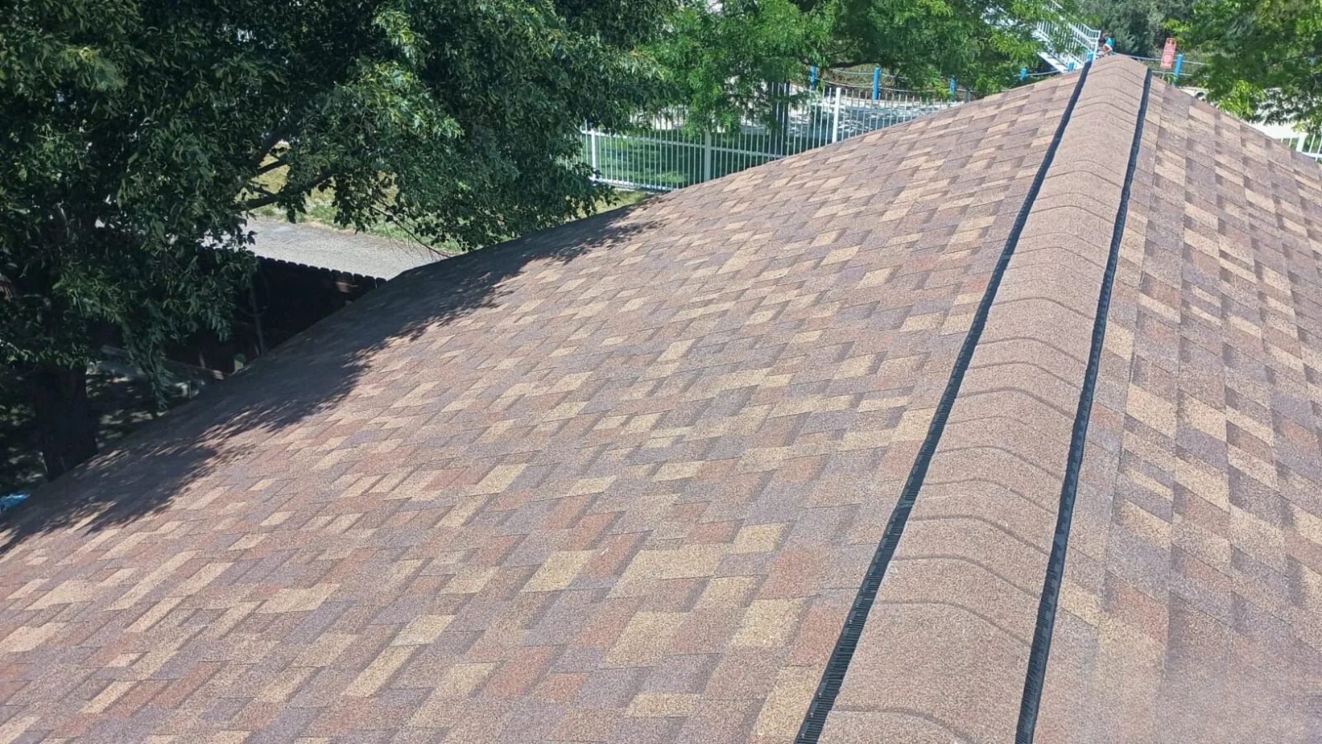 A roof with brown shingles in a residential area. The roof is partially shaded by a large tree, and a white fence is visible in the background.