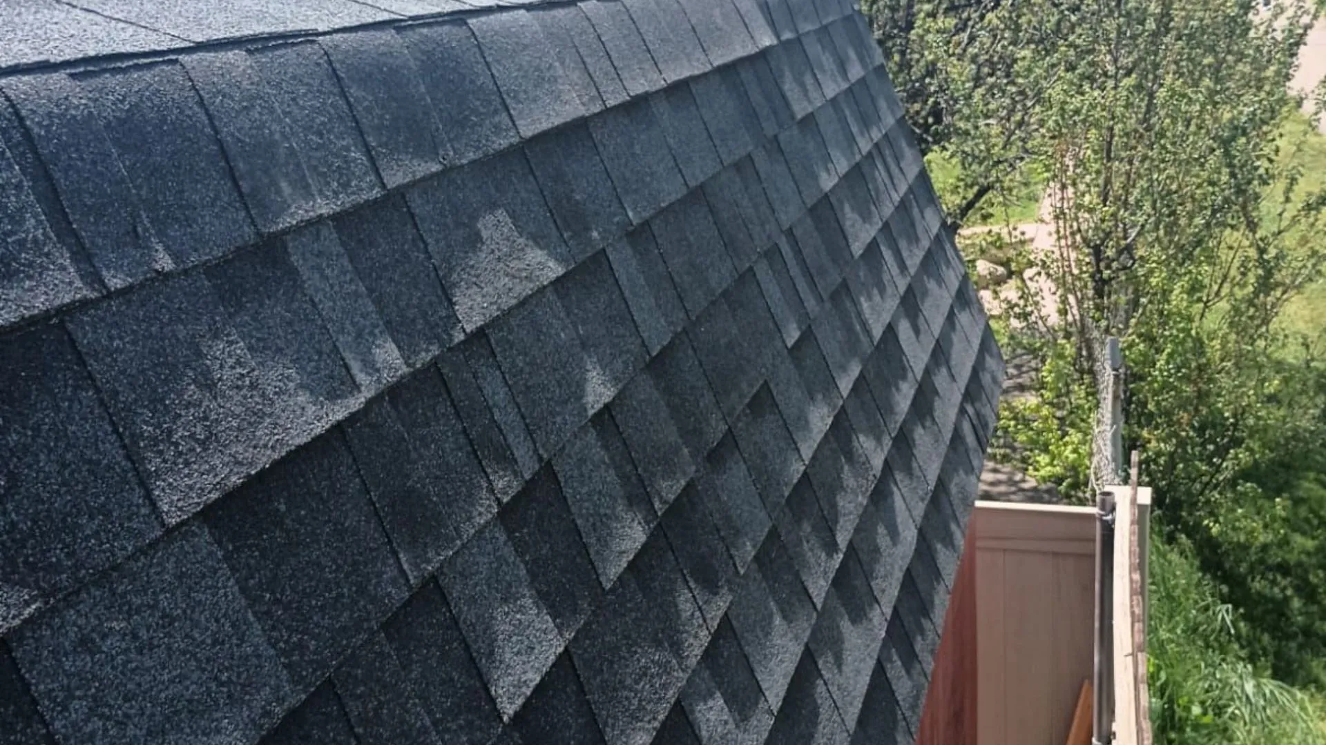 Close-up view of a steep roof with dark gray shingles. The background shows some green trees and a fence.