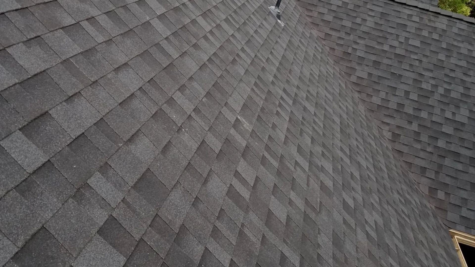 A roof with gray shingles, featuring a smooth and even shingle pattern. The background shows more residential roofs and greenery.