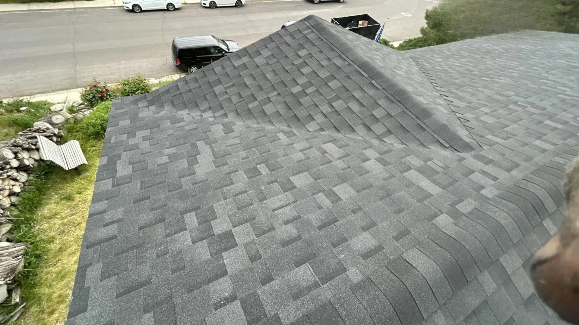 A roof with dark gray shingles viewed from an elevated perspective, showing a well-maintained shingle pattern and a quiet street with cars parked.