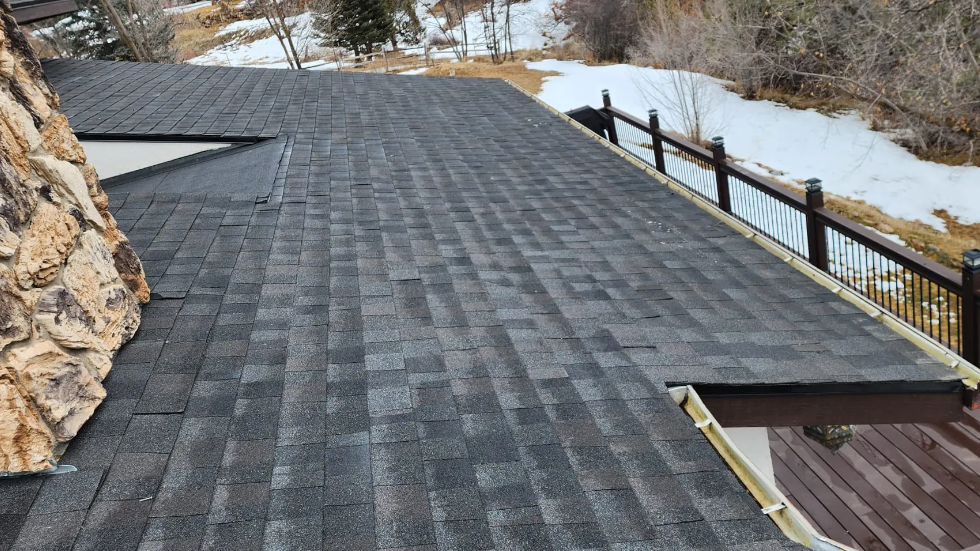 A roof with dark shingles next to a stone chimney, overlooking a snowy landscape and a deck with a railing.