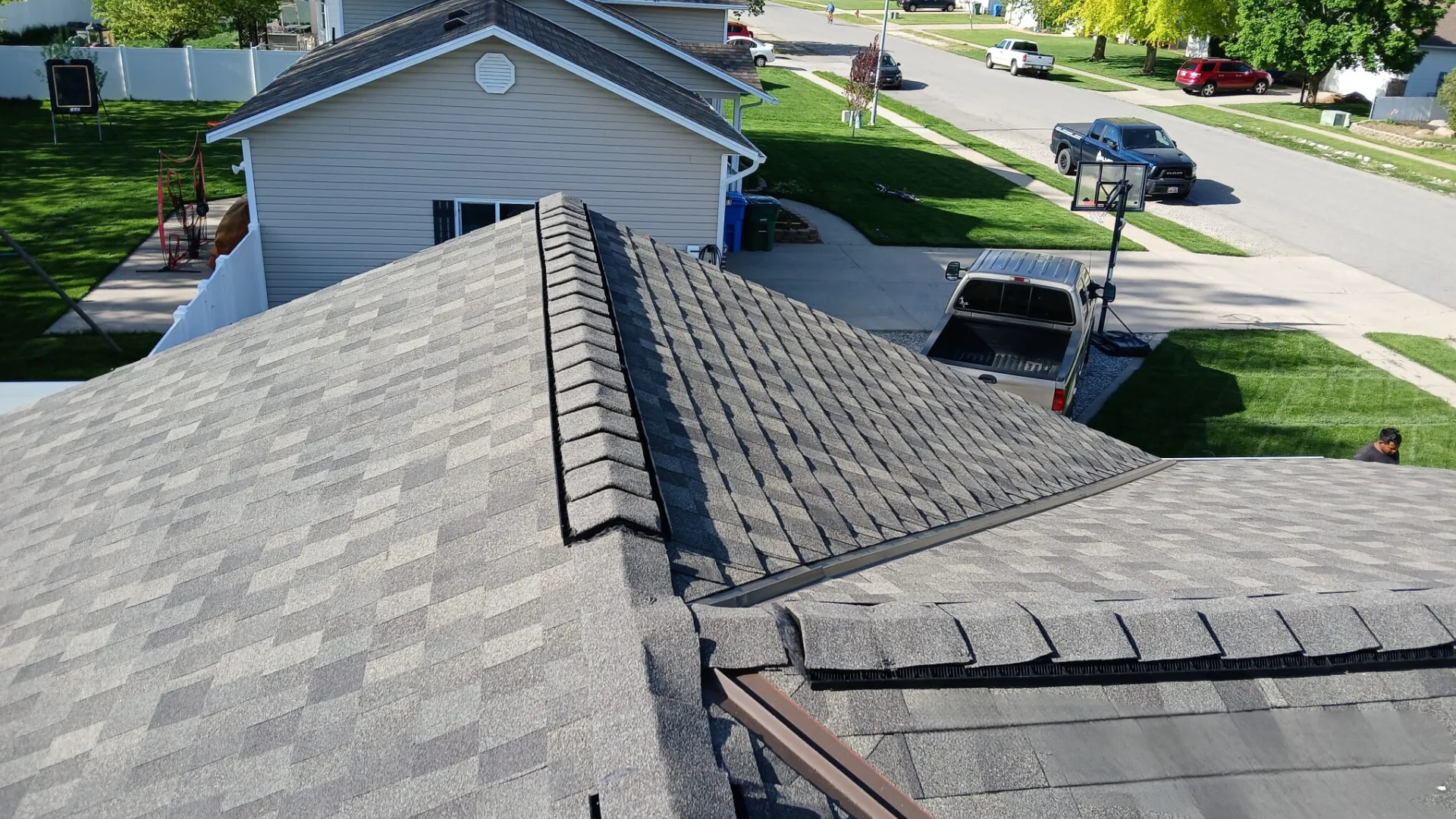 View of a gray shingled roof from above, showing a well-maintained residential area with houses, driveways, and green lawns.