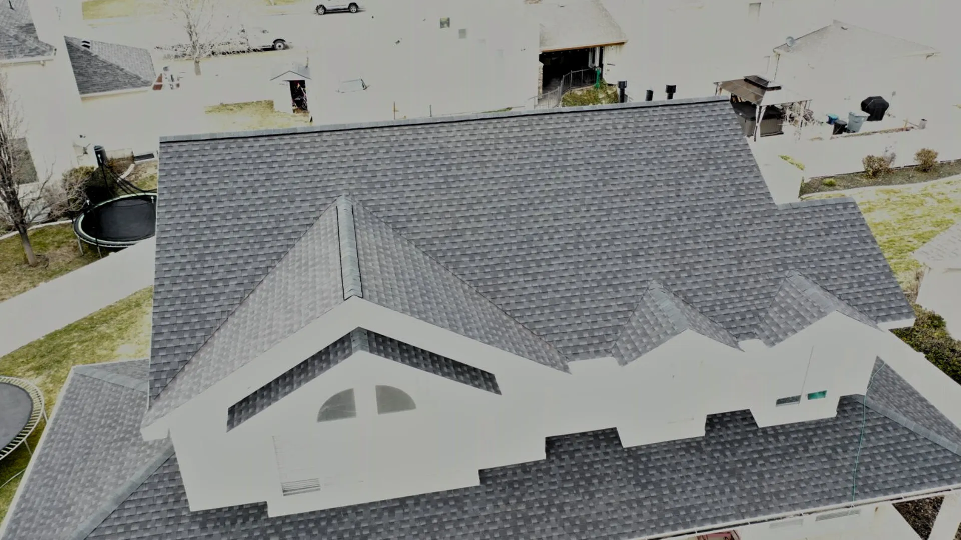 Aerial view of a dark shingled roof with various roof structures and chimneys, set in a residential area with snow on the ground.