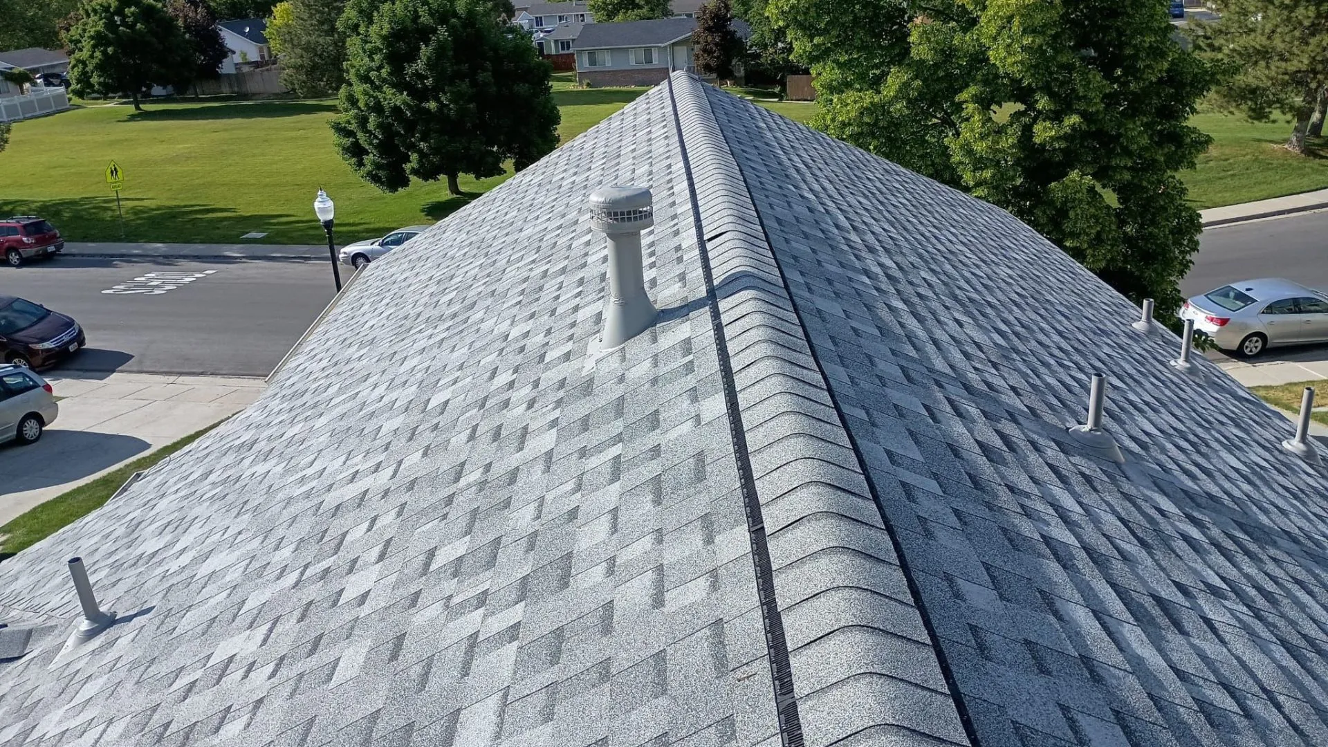 A roof with light gray shingles viewed from above, set in a suburban neighborhood with a street and parked cars visible.