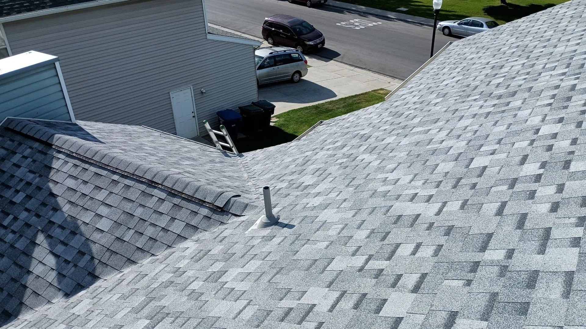 Close-up of a brown shingled roof with a detailed view of the shingle pattern and a vent pipe. The background includes a green backyard.