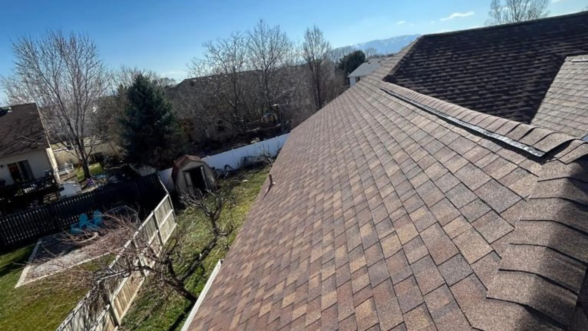 View of a long, brown shingled roof with a clear blue sky in the background. Trees without leaves are visible along with other houses and backyards.